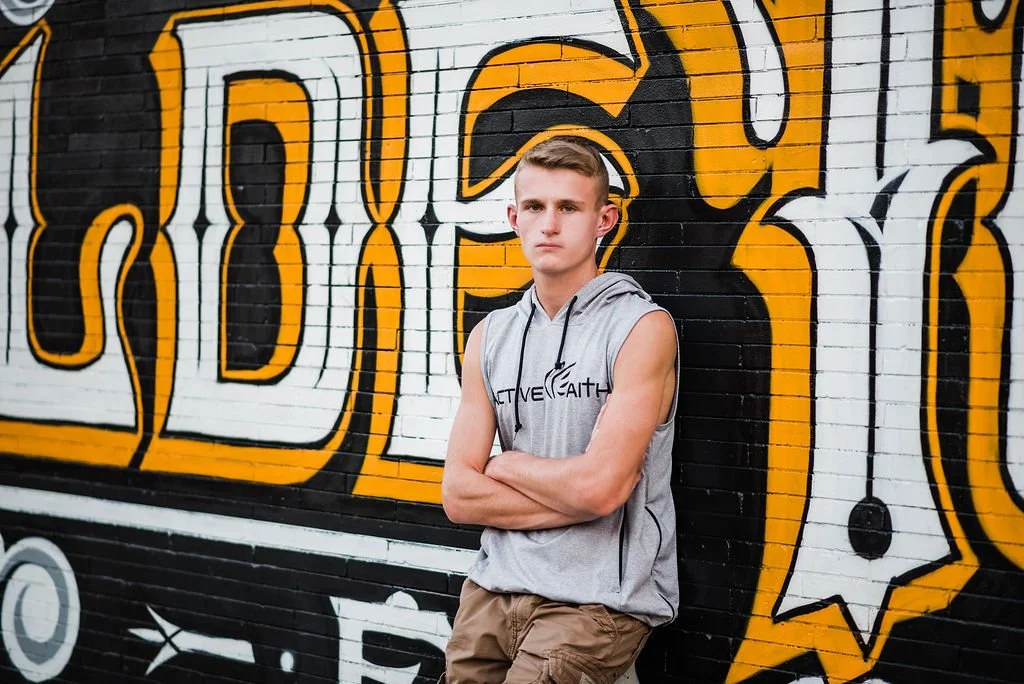 A young man with short blond hair wearing a sleeveless gray hoodie with the words 'Active Faith' printed on it, standing with arms crossed against a black brick wall with large colorful graffiti art.