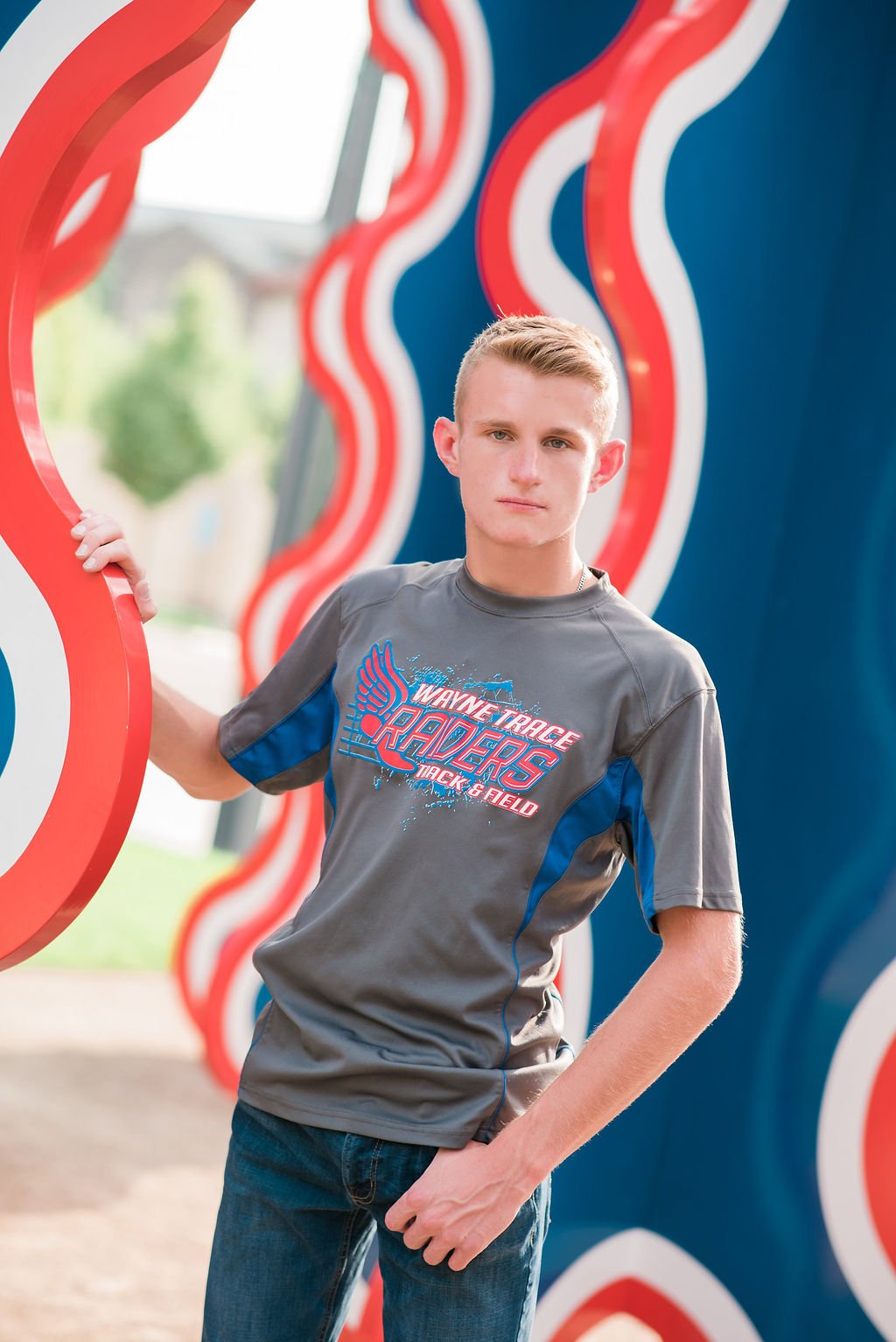 A young man with short blonde hair wearing a gray and blue sports T-shirt standing outdoors next to a colorful wavy structure, looking at the camera with a serious expression.