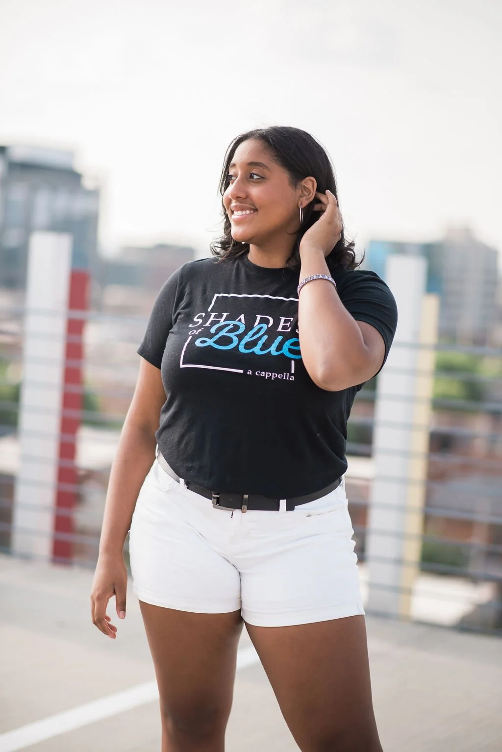 Young woman standing on a rooftop, wearing a black t-shirt with the text "Shades of Blue" and white shorts, with city buildings in the background.