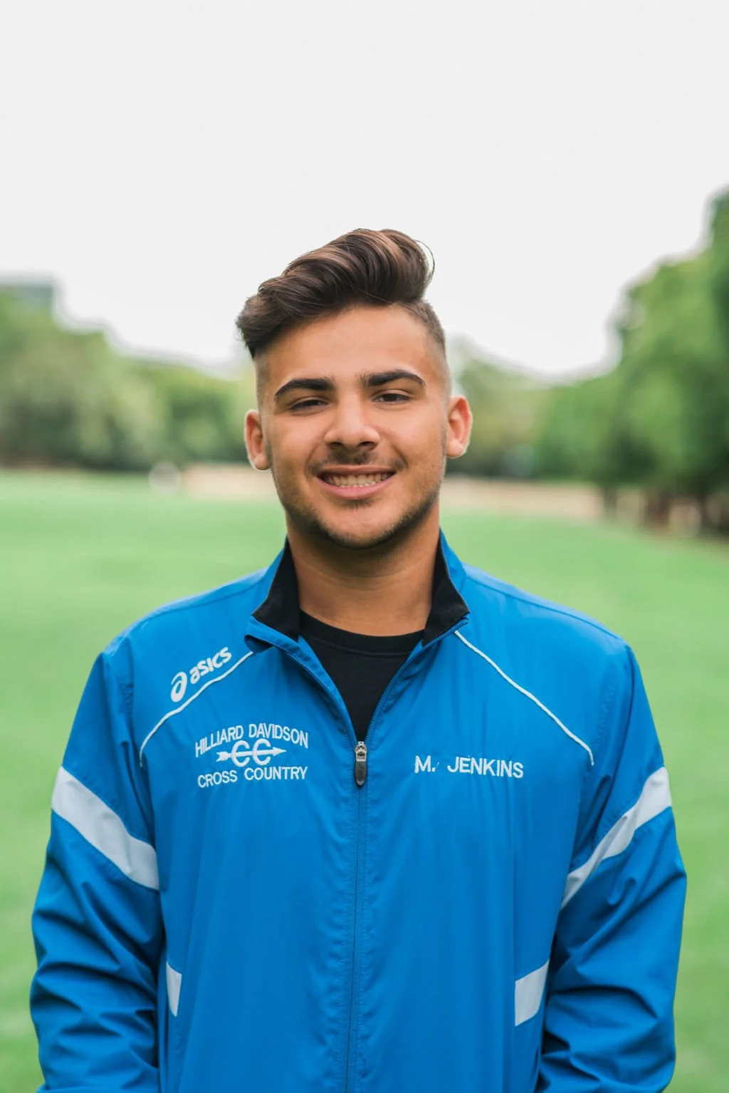 Young man smiling outdoors, wearing a blue sports jacket with 'Hilliard Davidson Cross Country' and his name embroidered on it, in a park with green trees in the background.