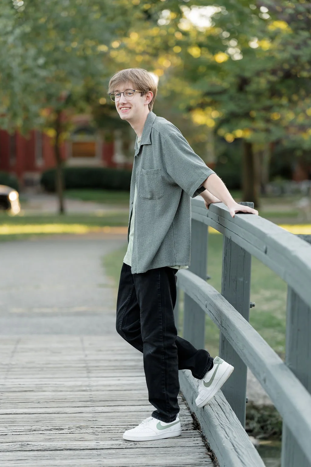 A young man with glasses wearing a gray checkered shirt, black pants, and white Nike sneakers, standing on a wooden bridge outdoors, leaning on the railing, with trees and houses in the background during late afternoon.