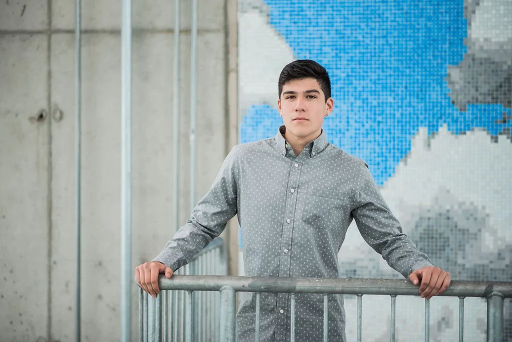Young man with dark hair and fair skin standing behind a metal railing indoors, with a pixelated map of the United States in blue, gray, and white in the background.