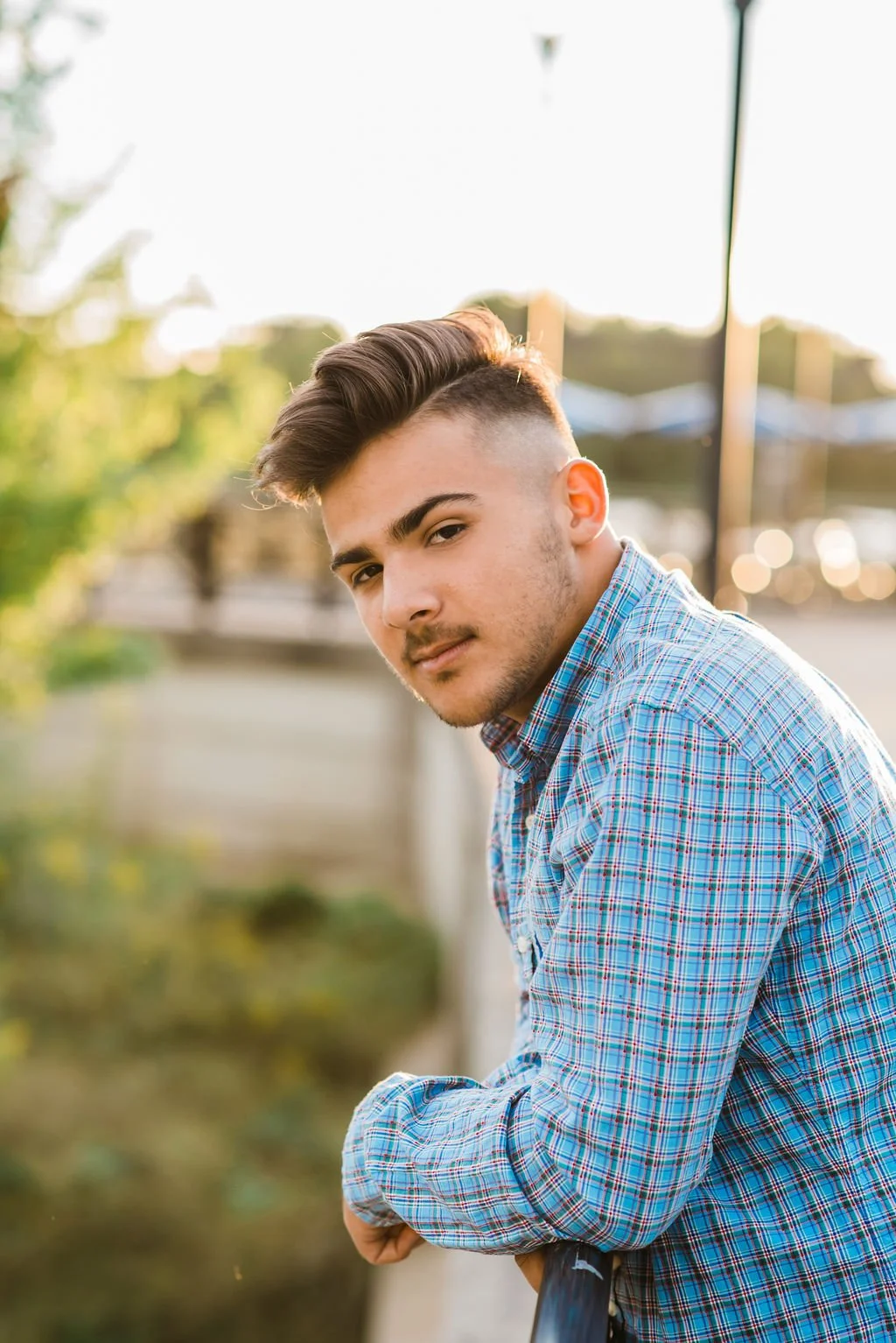 A young man leaning on a railing outdoors during sunset, wearing a blue plaid shirt and looking into the camera.