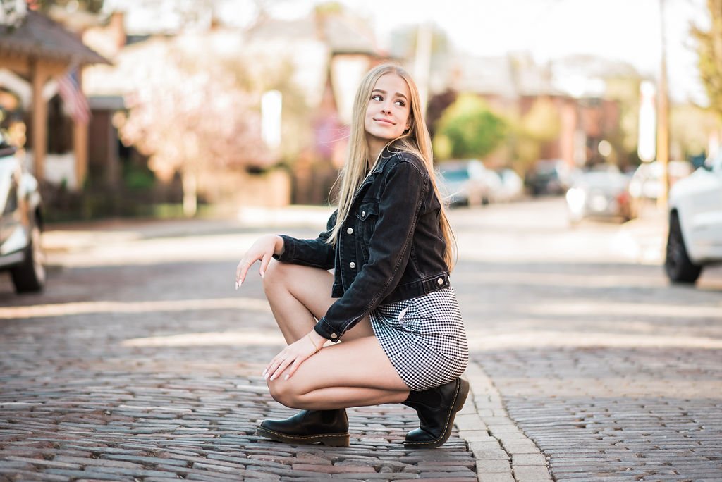 Young woman with long blonde hair in a black denim jacket and checkered shorts, squatting on a cobblestone street in a residential neighborhood, with cars and colorful houses in the background.