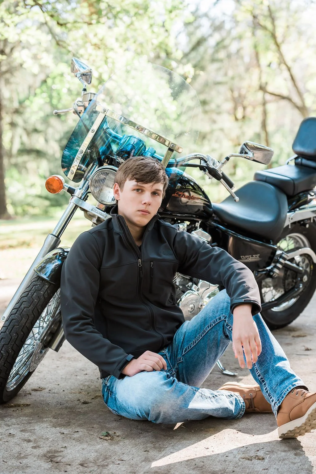 A teenage boy sitting on the ground and leaning against a black motorcycle parked outdoors in a wooded area.