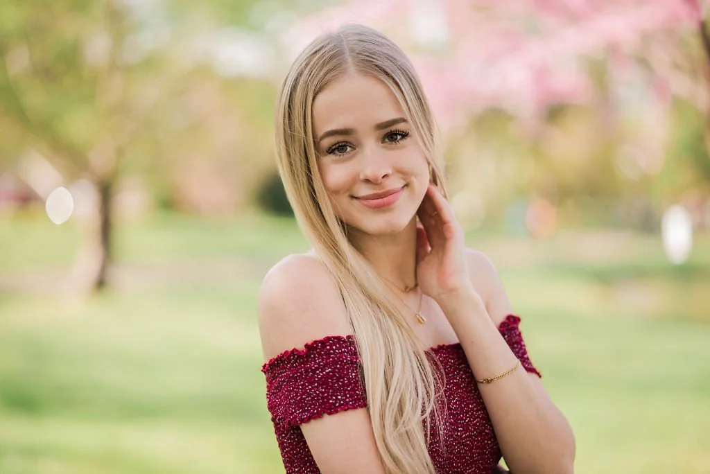 A young woman with long blonde hair wearing an off-the-shoulder red dress outdoors, smiling at the camera with a blurred natural background.