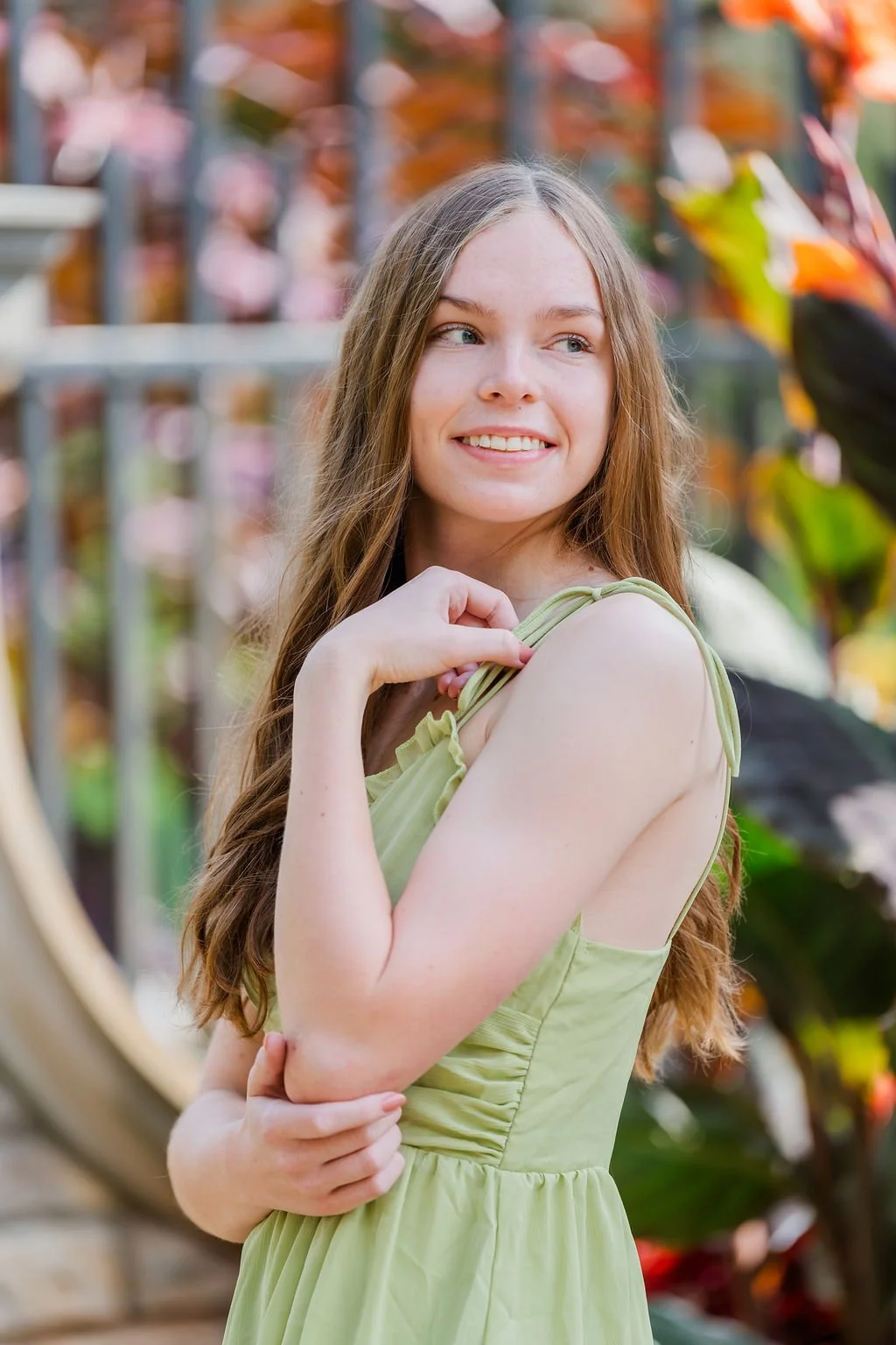 Young woman with long, light brown hair wearing a light green dress, smiling and looking to the side, outdoors with colorful foliage in the background.