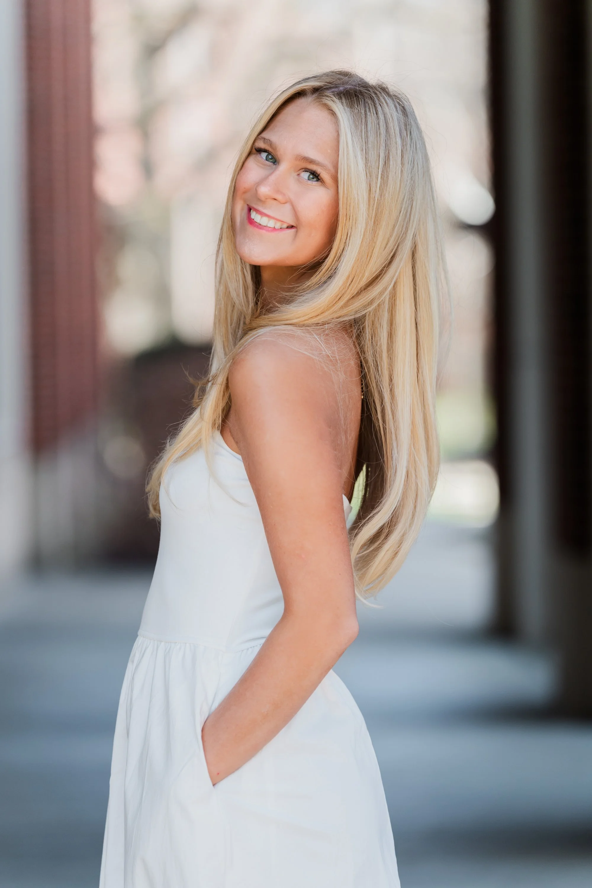 A young woman with long blonde hair wearing a white dress standing outdoors with her hands in her pockets, smiling at the camera.