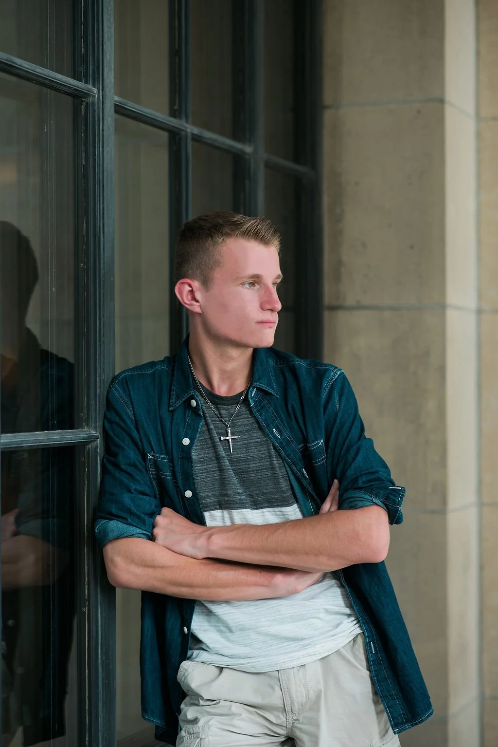 A young man with short hair, wearing a cross necklace, a striped t-shirt, a denim shirt, and khaki pants, standing with crossed arms, looking to his right against a building with large windows and stone walls.