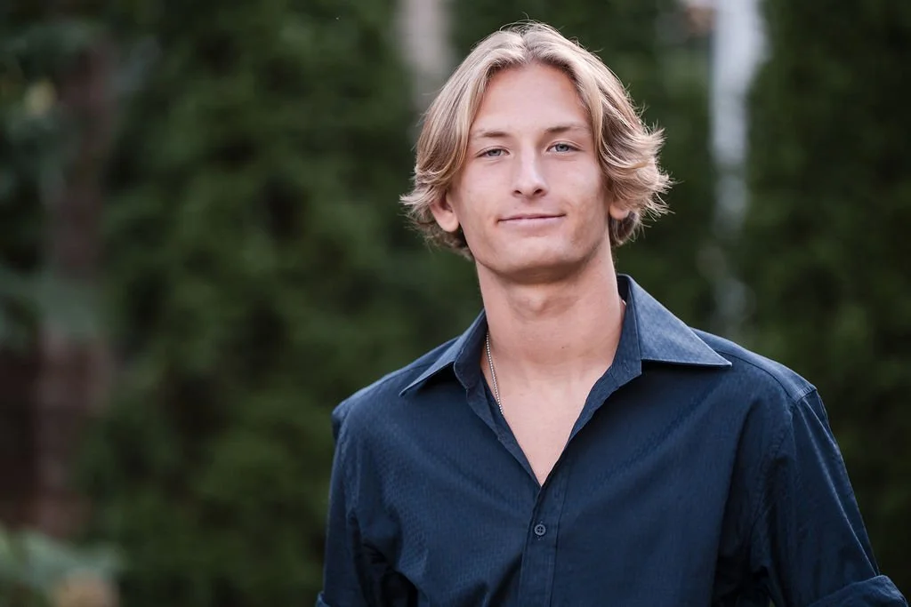 A young man with blond, wavy hair and light skin, wearing a dark blue shirt, standing outdoors with a background of blurred greenery.