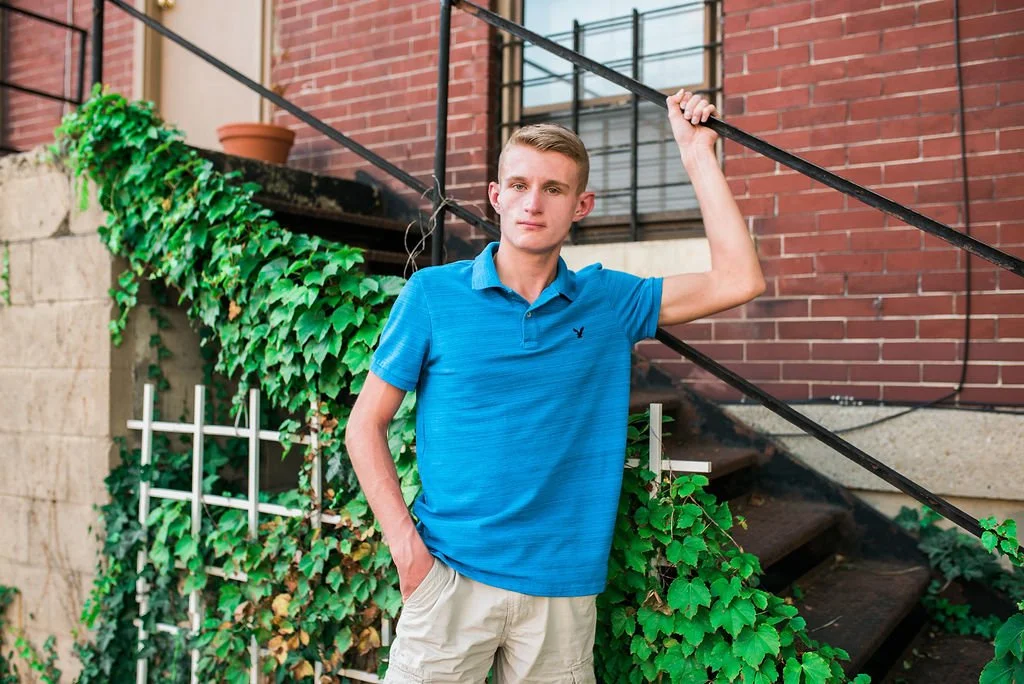 A young man in a blue polo shirt and khaki shorts standing outdoors, holding onto a black handrail on a staircase next to a brick building with green ivy growing on a lattice.