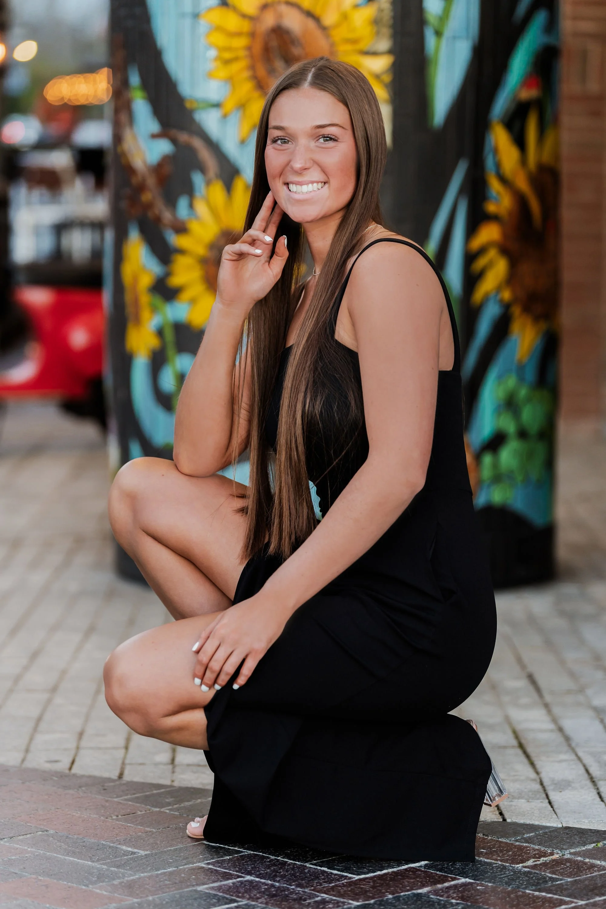 A young woman with long brown hair kneeling on a sidewalk, smiling at the camera with a colorful mural of sunflowers behind her.