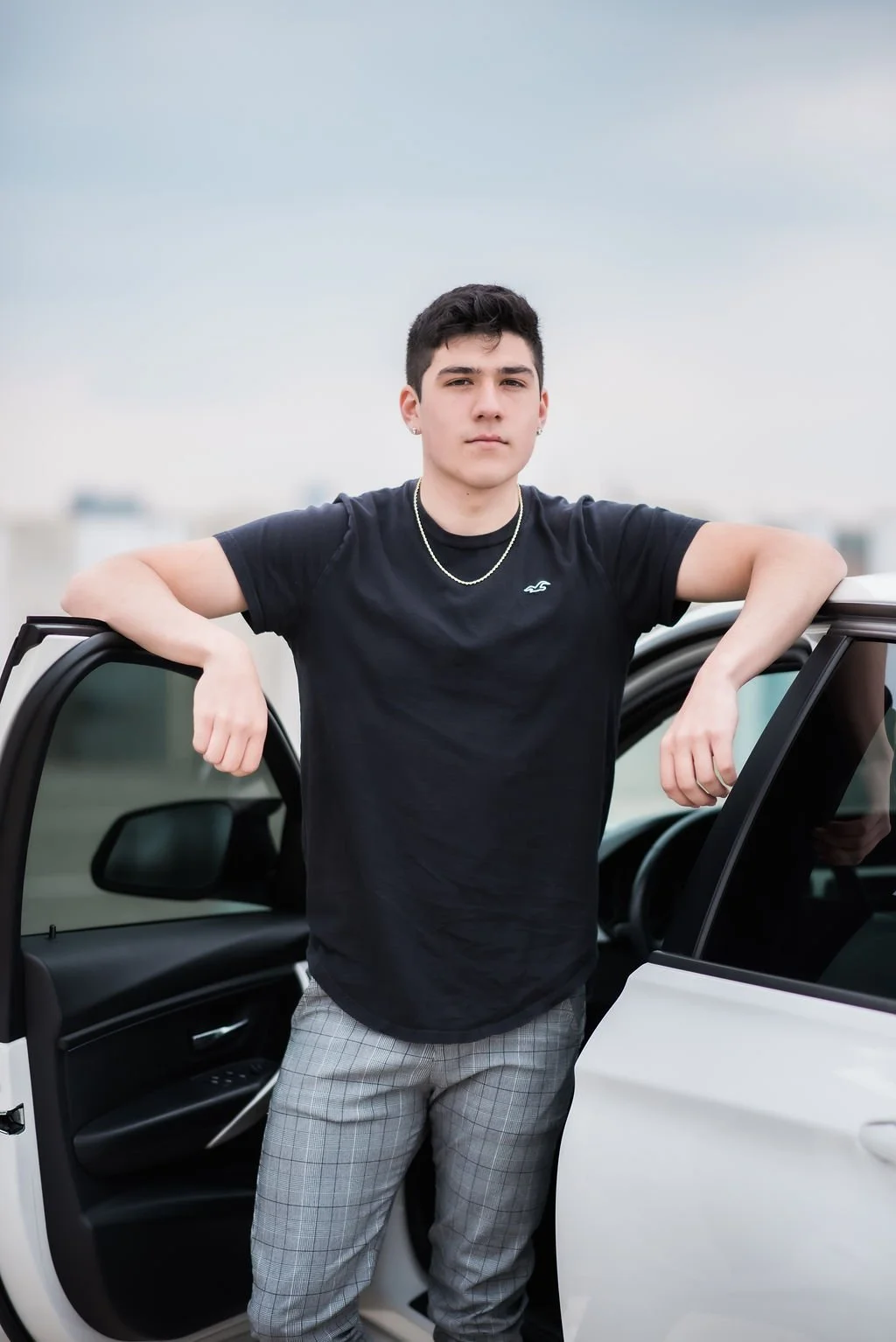 A young man standing beside a white car with the driver's door open, outdoors on a cloudy day.