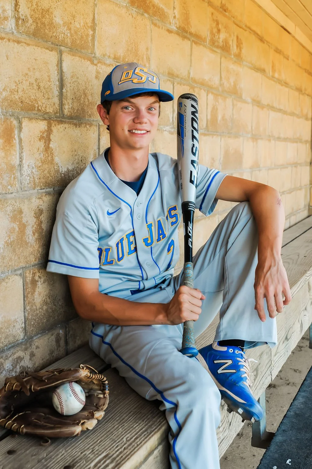 A young baseball player in a light gray and blue uniform sitting on a wooden bench against a brick wall, holding a baseball bat, with a baseball glove and ball nearby.