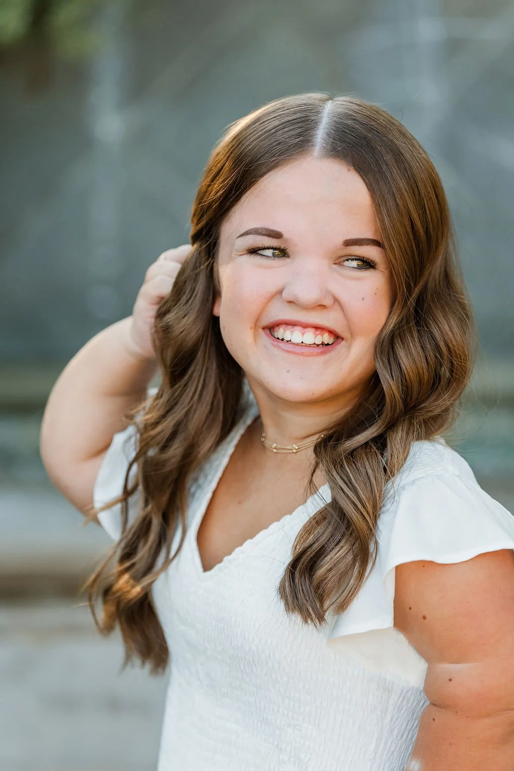 A smiling woman with long wavy brown hair and light skin, wearing a white top, posing outdoors in front of a blurred background of water and trees.