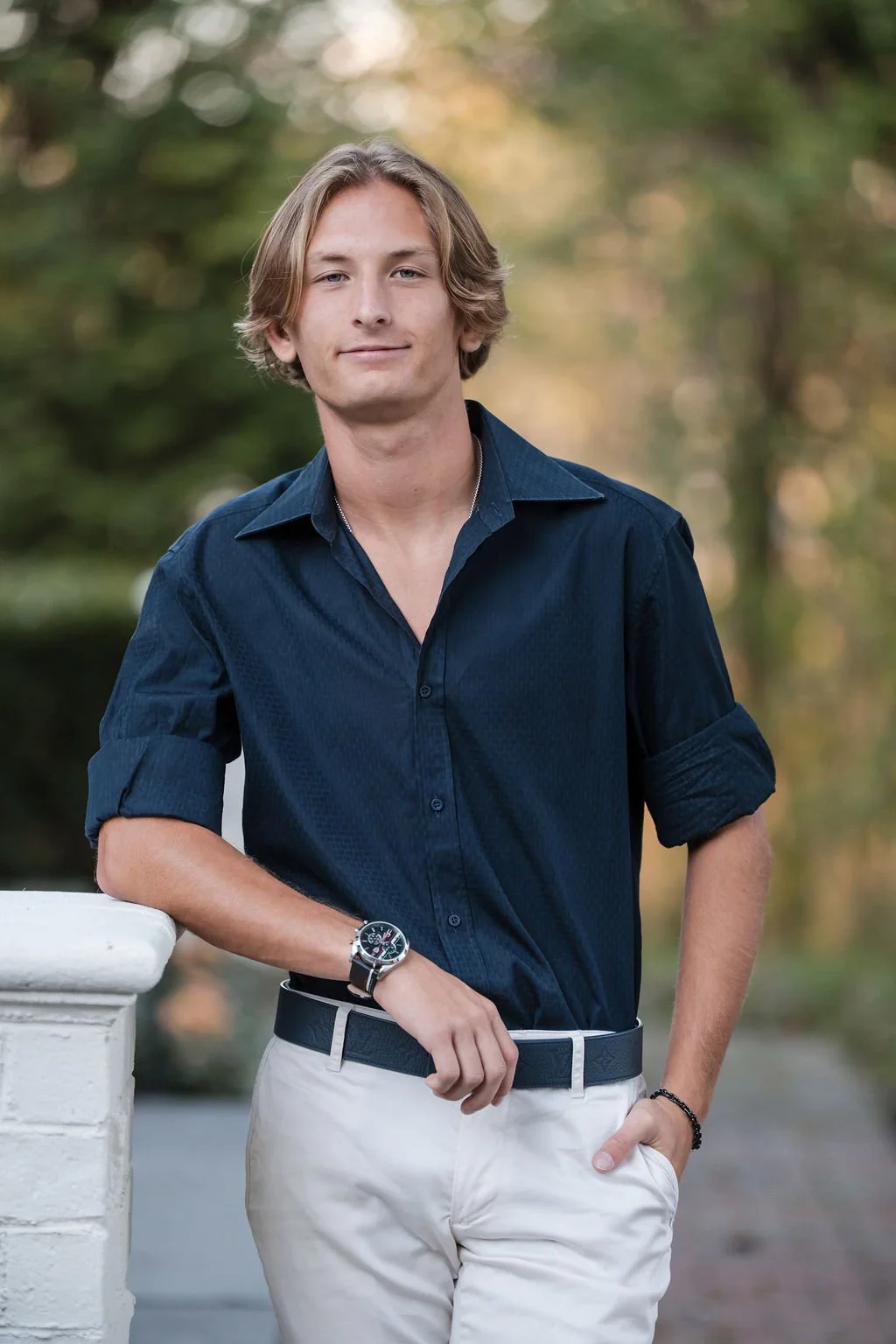 A young man with shoulder-length blond hair, wearing a dark blue button-up shirt with rolled-up sleeves, beige pants, a watch on his left wrist, and a bracelet on his right wrist, standing outdoors with a blurred background of trees and greenery.