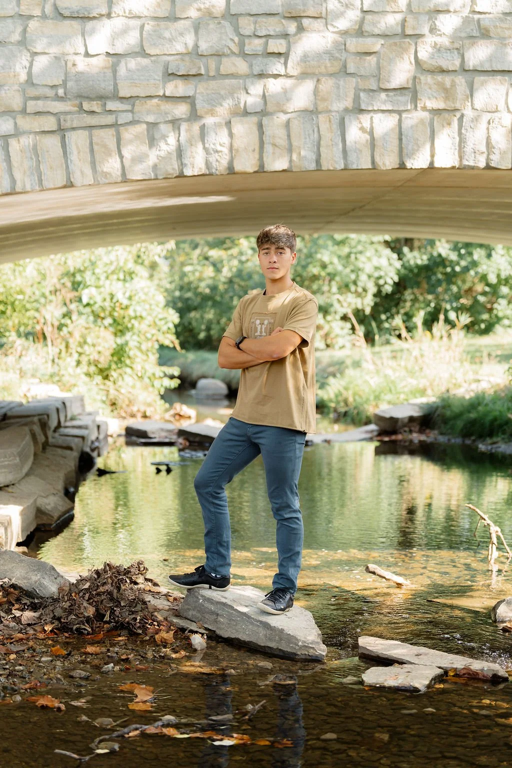 A young man standing on a rock in a shallow creek underneath a stone bridge, with lush green trees in the background.