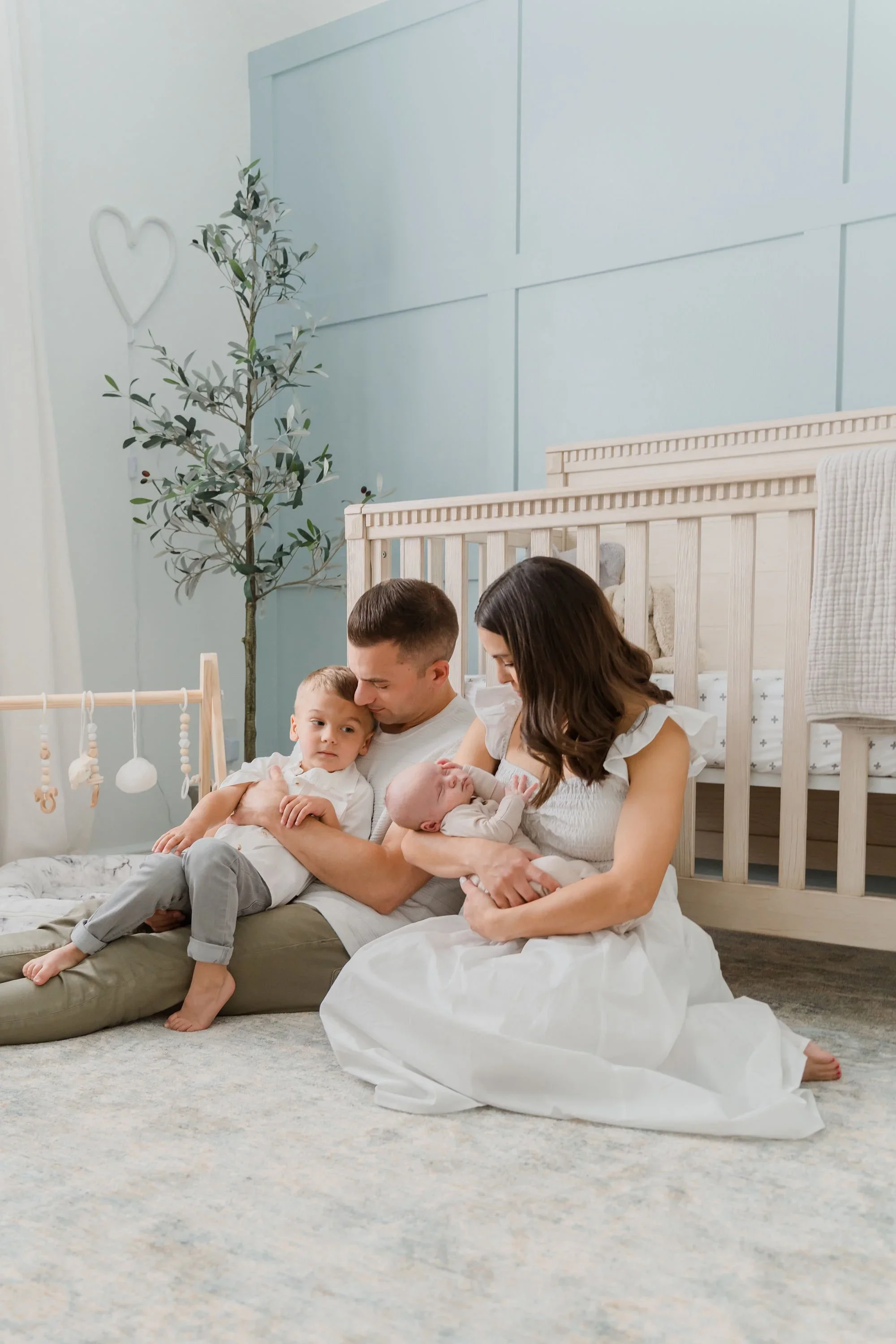 A family of four sitting on a carpet in a nursery, with the mother holding a newborn and the father holding an older child, all sharing a tender moment.