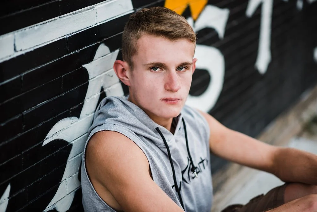 A young man with short, light brown hair and light skin, wearing a sleeveless hoodie, sitting against a black and white graffiti wall and looking at the camera.