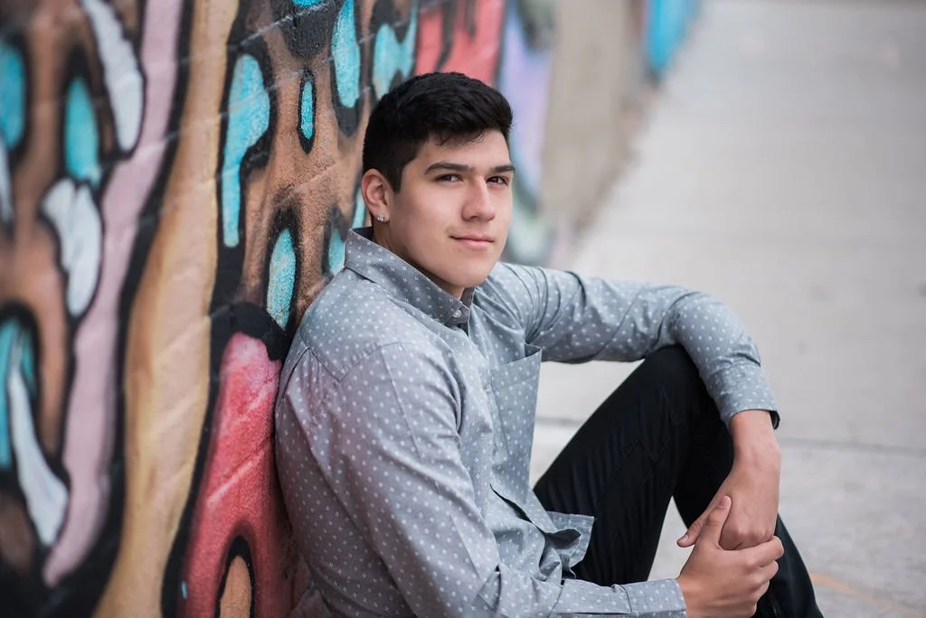 A young man sitting on the sidewalk, leaning against a colorful graffiti wall, looking at the camera with a slight smile.