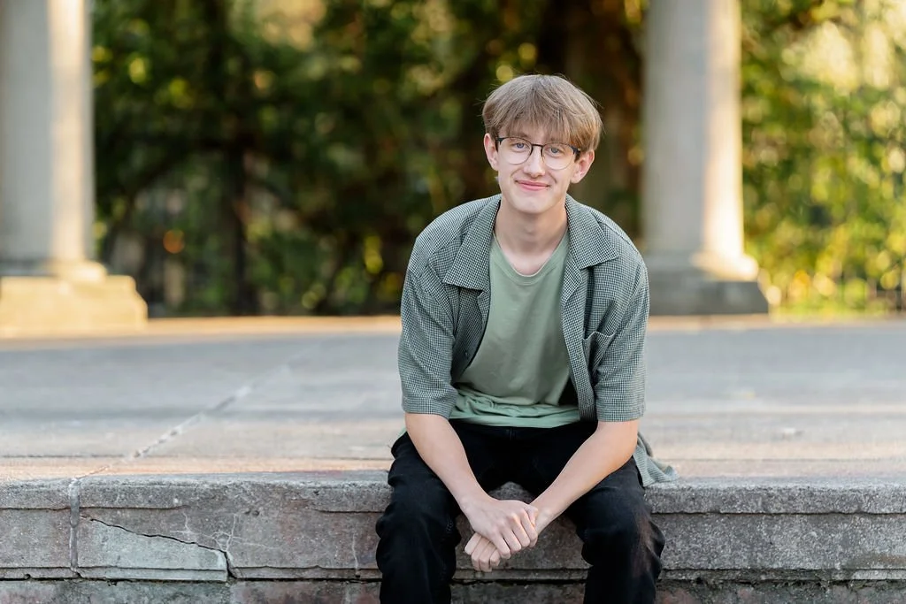 A young man with glasses sits on a stone ledge outdoors, smiling with a background of green trees.
