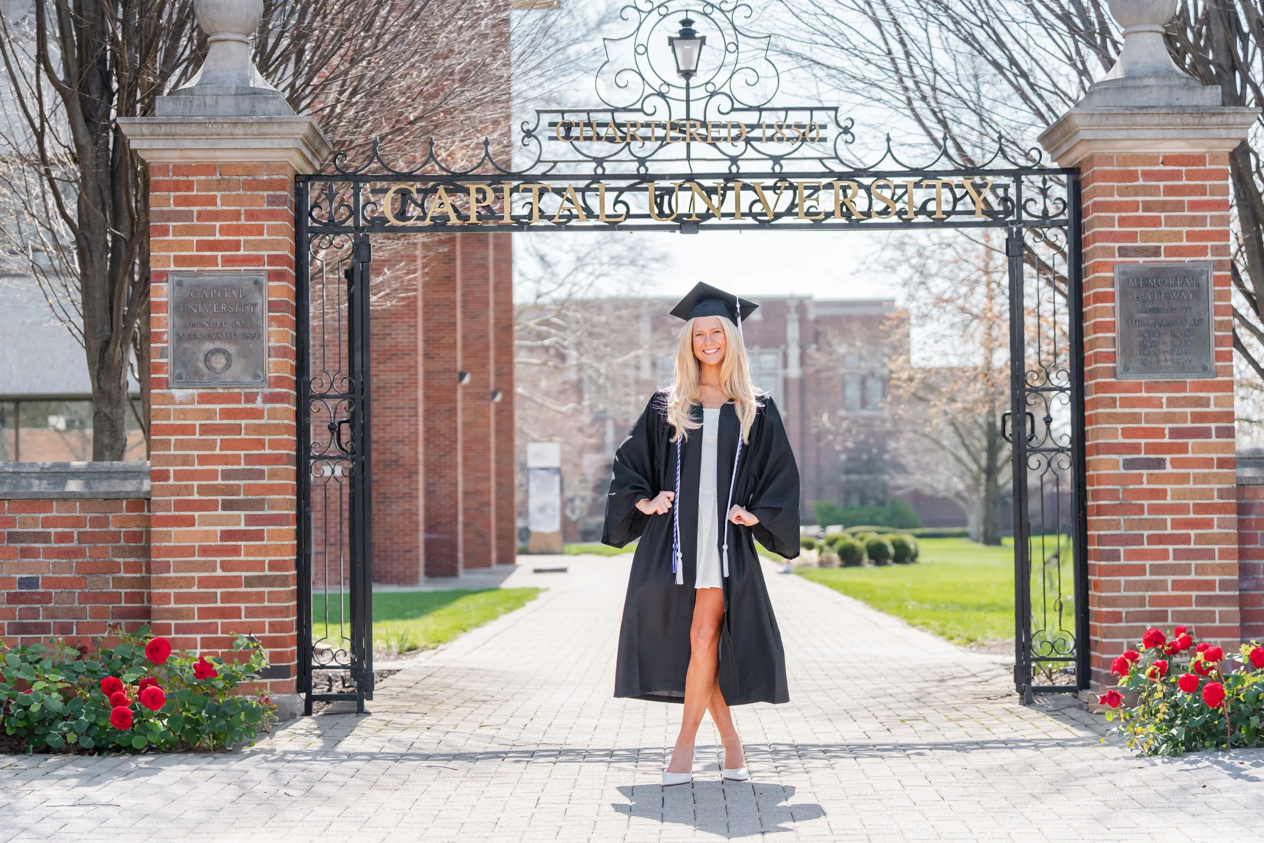 A young woman in a graduation cap and gown standing with a smile under an ornate metal archway labeled 'CAPITAL UNIVERSITY.' She is outdoors on a sunny day with brick buildings and leafless trees in the background.