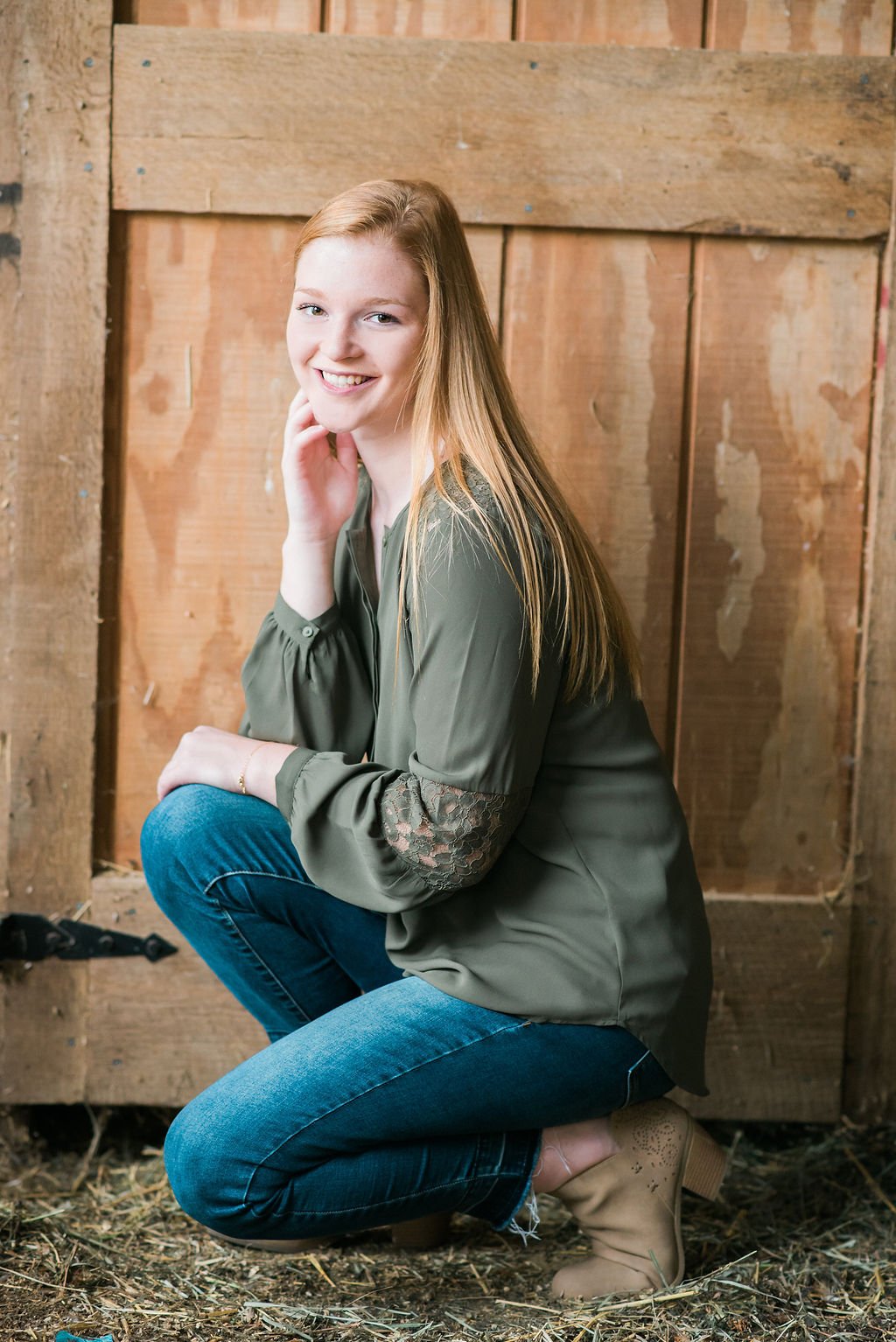 A young woman with long red hair poses inside a wooden barn, wearing a green blouse with lace sleeves, blue jeans, and tan boots, kneeling on the dirt floor with a cheerful expression.