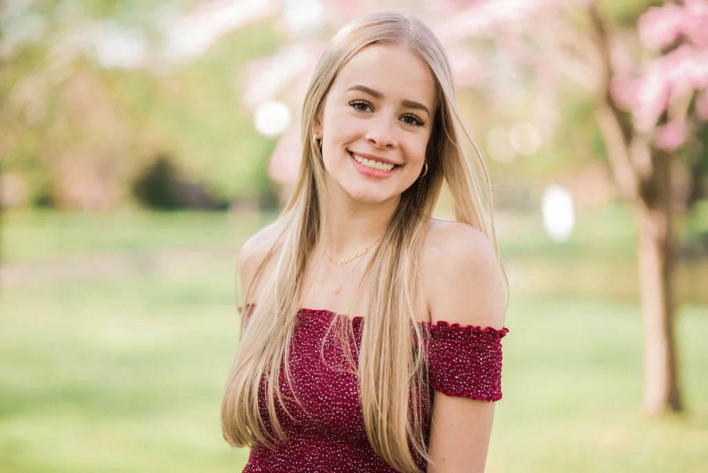A young woman with long blonde hair smiling outdoors during spring, wearing a red off-the-shoulder dress with sparkles.