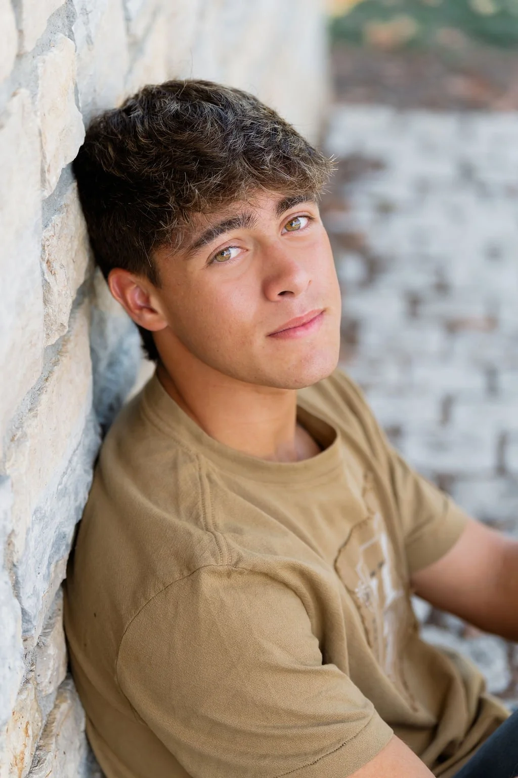 Young man with brown hair and light eyes, sitting against a stone wall, wearing a tan T-shirt, looking at the camera.