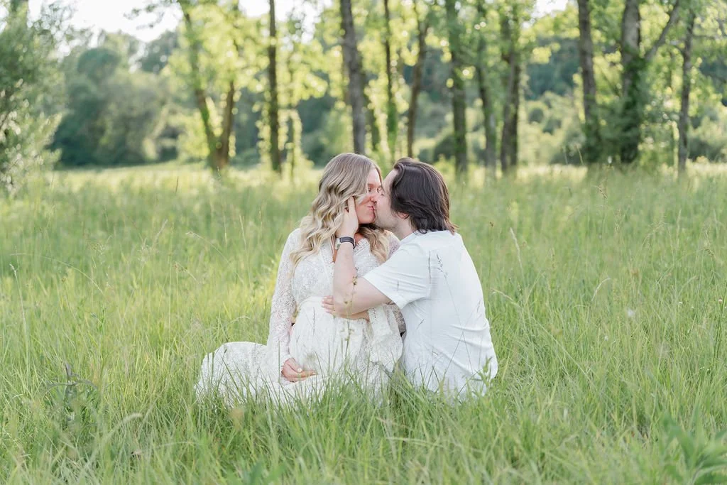 A couple sitting in a grassy field, sharing a kiss, surrounded by trees and greenery.