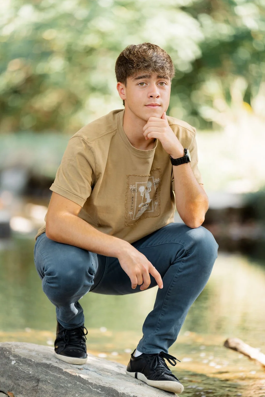 A young man crouching on a rock in a natural outdoor setting, with blurred green foliage and water in the background. He is dressed in a beige t-shirt, blue jeans, and black sneakers, and is resting his chin on his hand while looking at the camera.