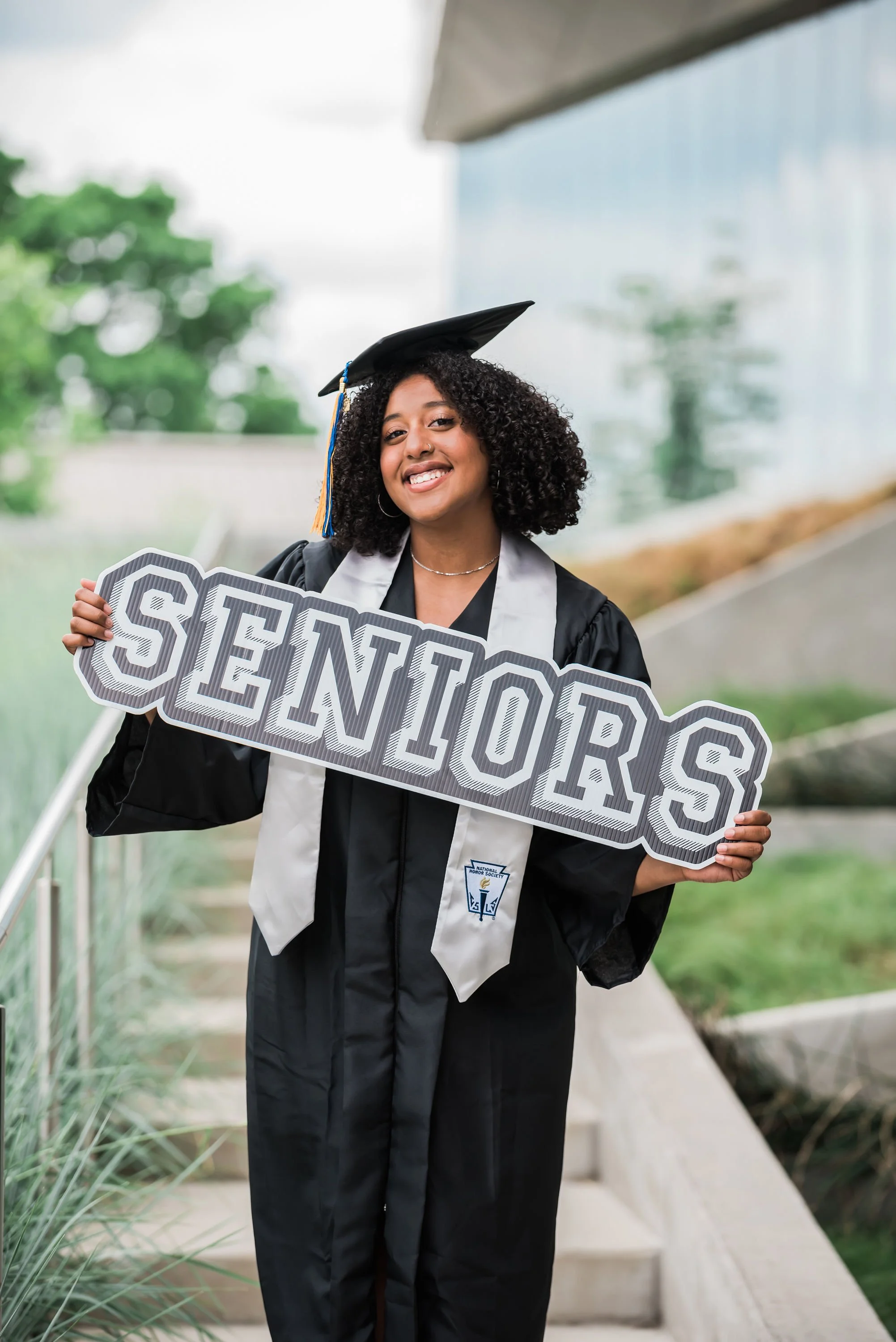 High school senior wearing graduation cap and gown holding a “Seniors” sign during an outdoor senior portrait session.