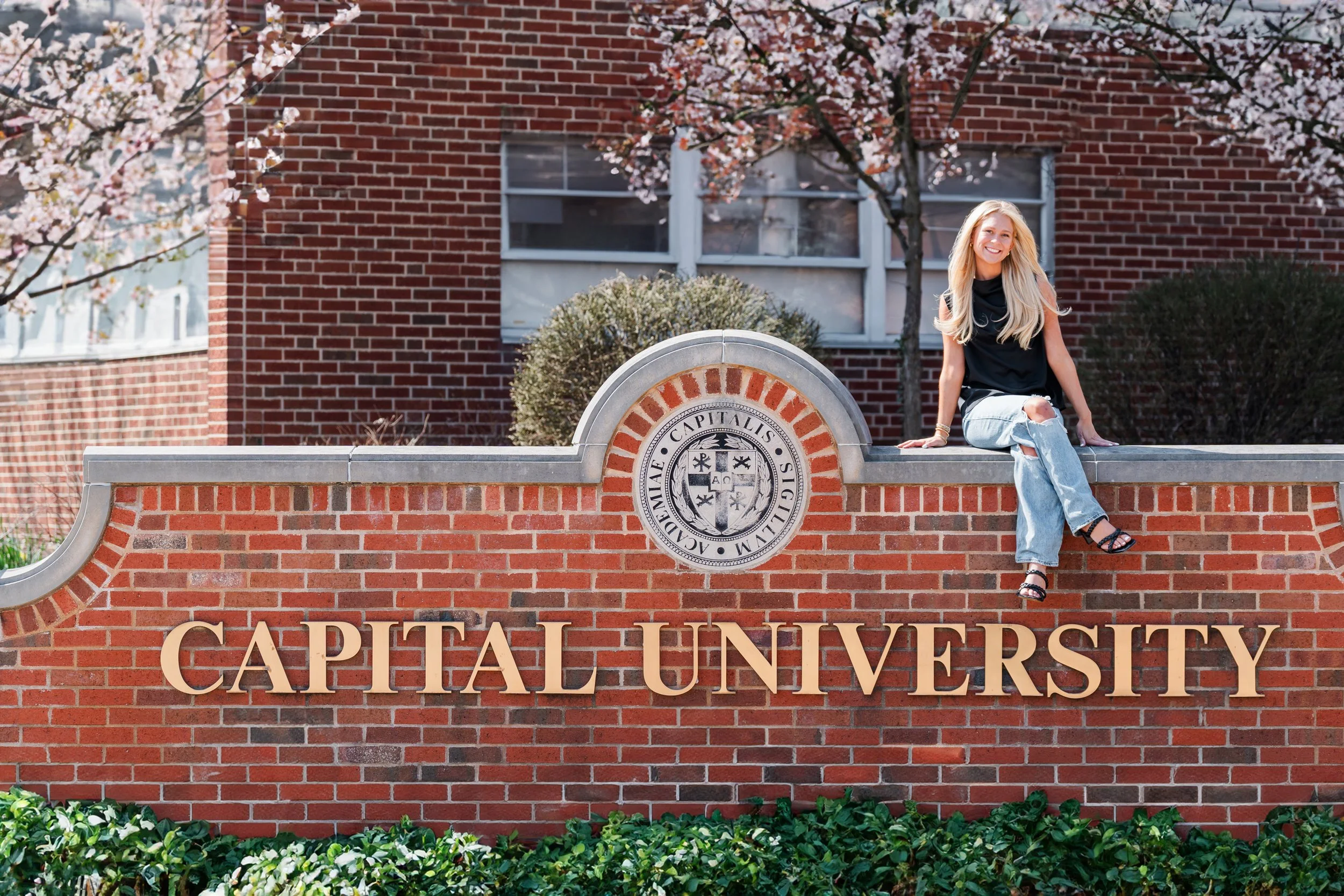 A young woman with long blonde hair, wearing a black sleeveless top and ripped blue jeans, sitting on a brick wall with gold letters spelling "Capital University" in front of a brick building with trees in bloom.