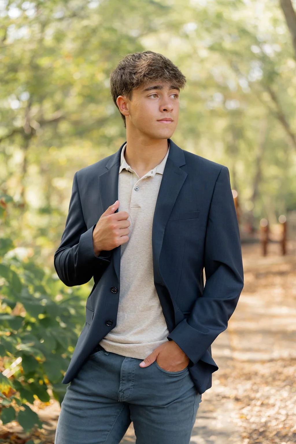 Young man in a dark blazer and light-colored polo shirt standing outdoors on a tree-lined path with green leaves and sunlight filtering through.
