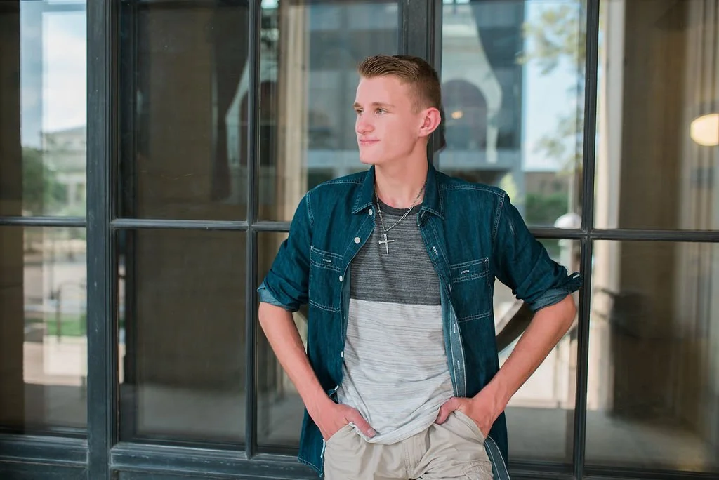 Young man with short hair, wearing a denim jacket over a gray and white striped t-shirt, standing with hands in pockets, looking to the side near glass doors.