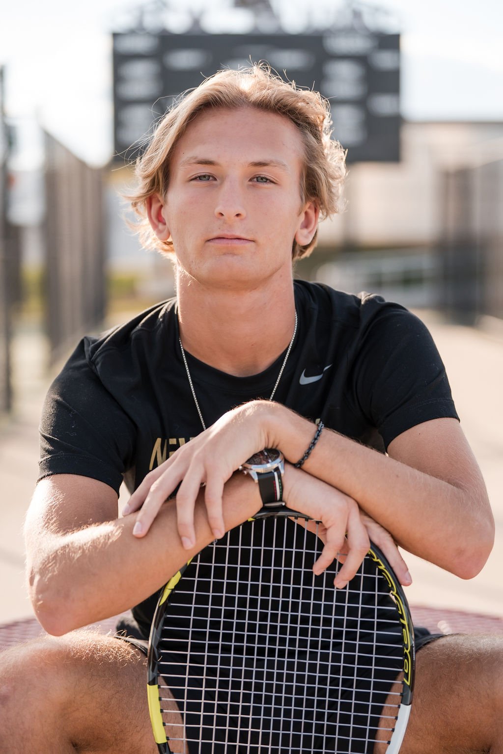 A young man with blond hair, wearing a black athletic shirt, sitting outdoors on a tennis court, holding a tennis racket, looking at the camera with a serious expression.