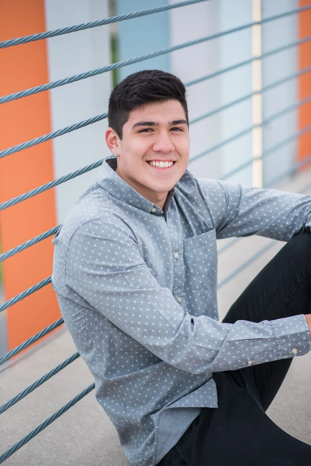 A young man with short dark hair, smiling, sitting on the ground near a metal railing with colorful vertical panels in the background.
