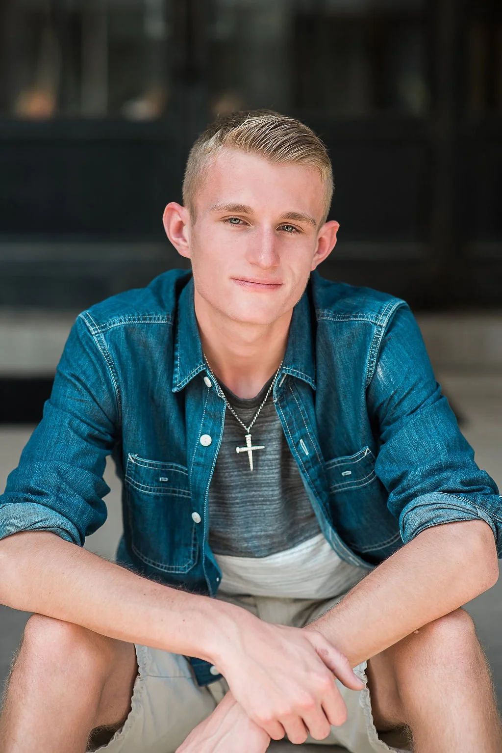A young man with short blond hair wearing a denim jacket, gray t-shirt, and a cross necklace, sitting outdoors with a neutral expression.