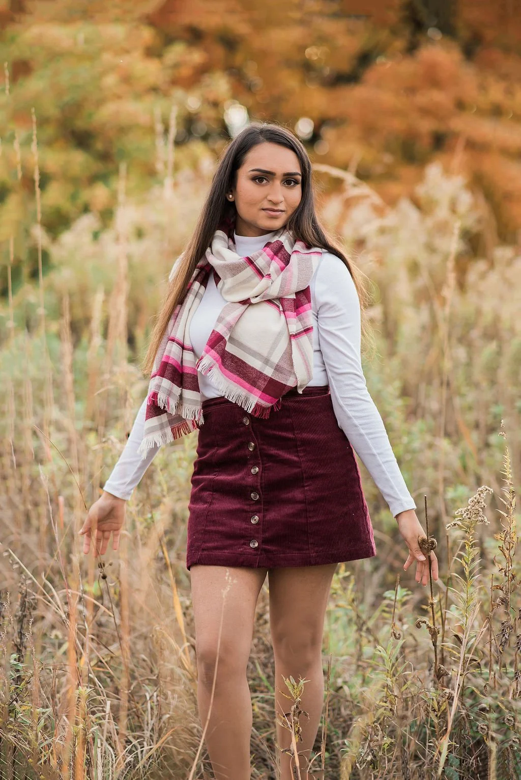 A woman standing in a field with autumn-colored trees in the background, wearing a white long sleeve shirt, maroon skirt with buttons, and a pink and beige plaid scarf, looking at the camera.