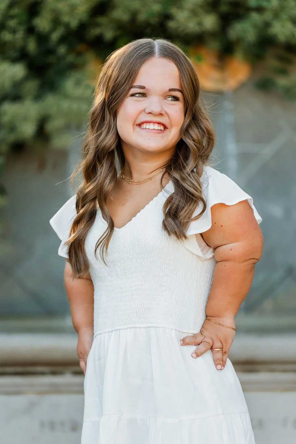 A smiling woman with long wavy brown hair wearing a white dress with puffed sleeves, standing outdoors with blurred trees and water in the background.