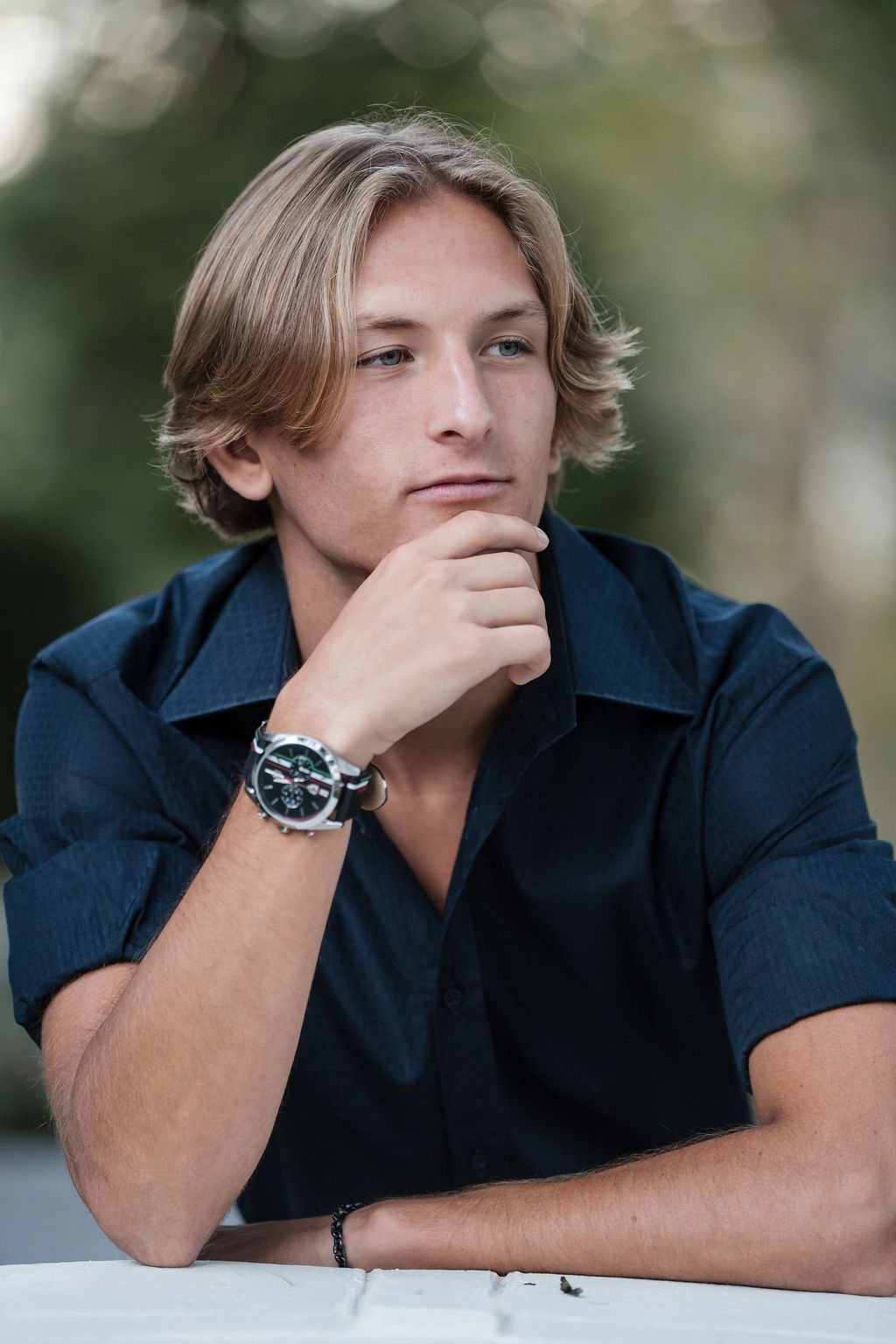 A young man with blonde hair and blue eyes, wearing a dark blue shirt and a wristwatch, sits outdoors with his hand resting on his chin, looking thoughtfully into the distance.