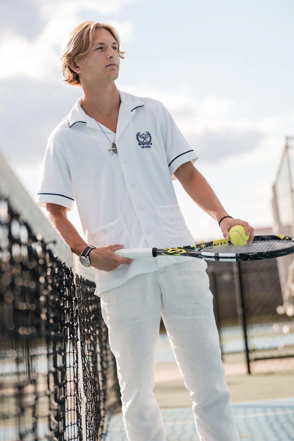A young man with blonde hair holding a tennis racket and a tennis ball, standing on a tennis court, dressed in white tennis attire with a cross necklace.