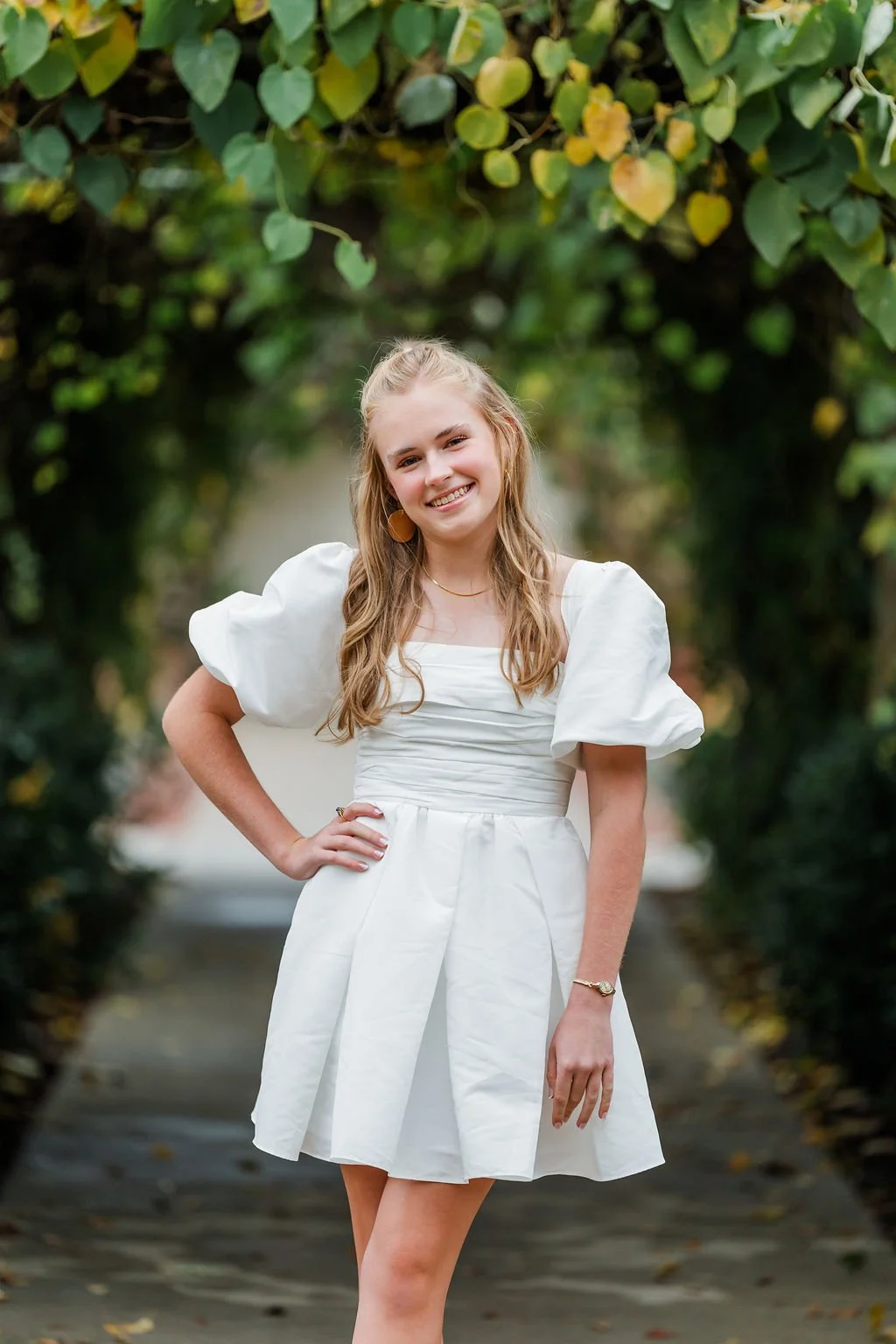 A young woman in a white dress posing outdoors on a path surrounded by green foliage.