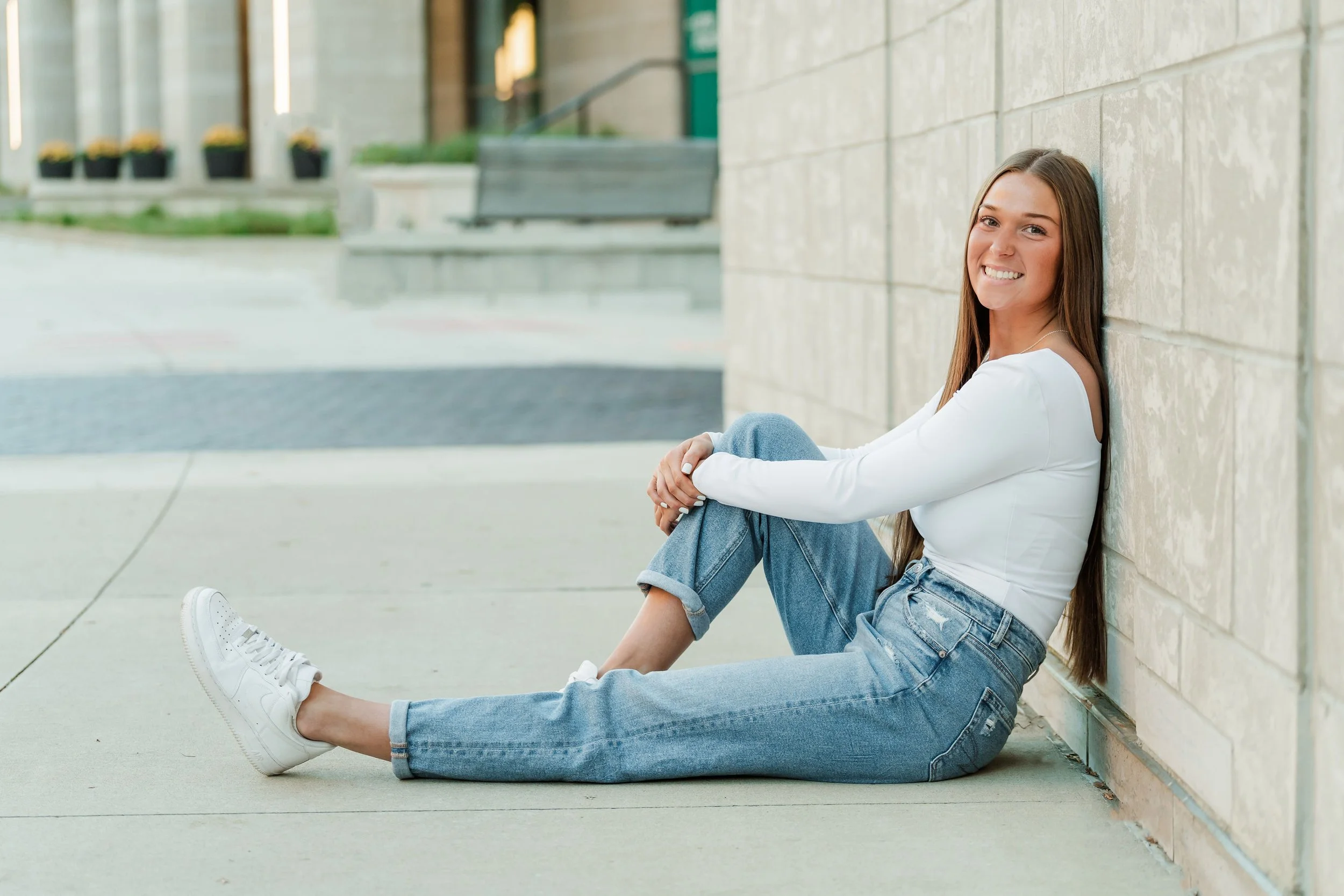 A young woman sitting on the sidewalk, leaning against a light-colored brick wall, smiling at the camera, wearing a white long-sleeve top, jeans, and white sneakers.