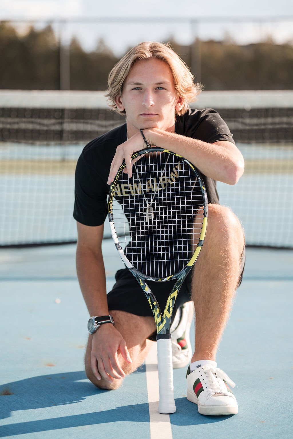 A young man kneeling on a tennis court, holding a tennis racket in front of him, with a serious expression, wearing a black t-shirt, shorts, and white sneakers.
