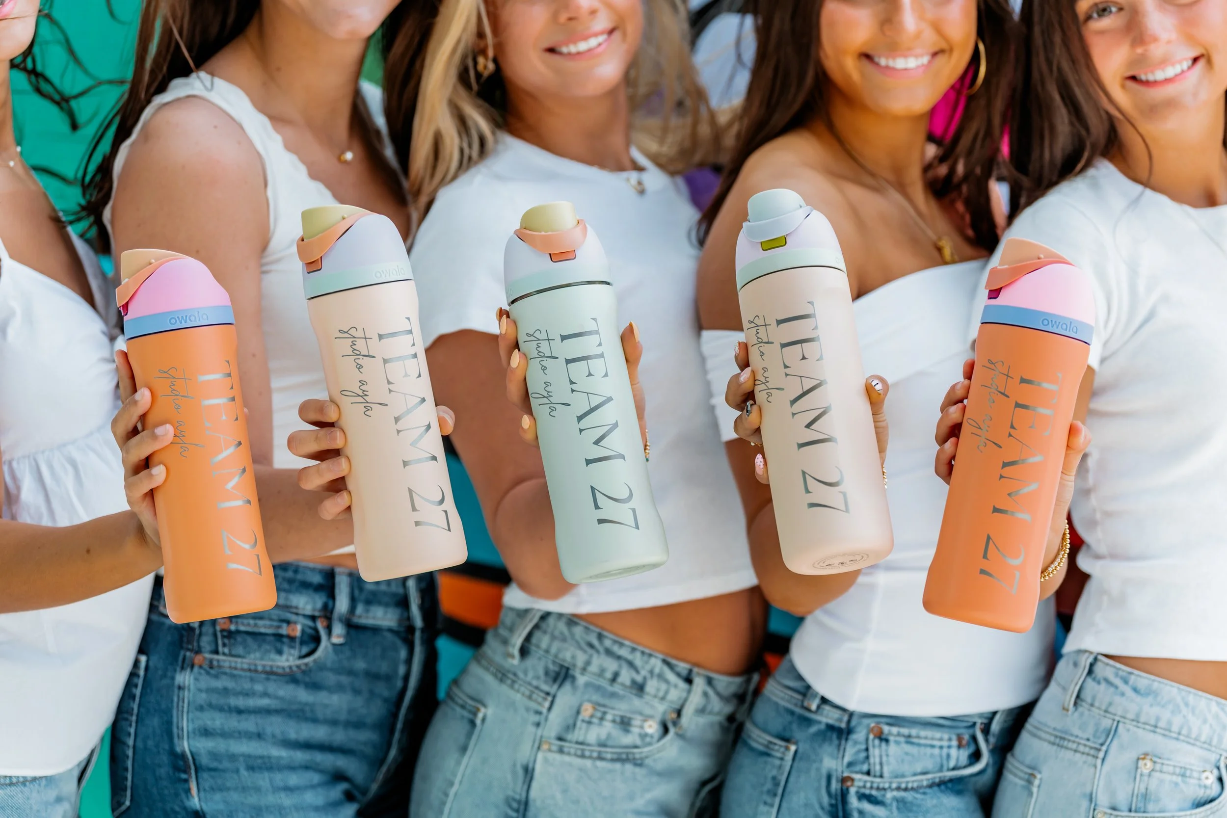 Group of women holding water bottles with 'Team 27' written on them, smiling and wearing white tops and jeans.