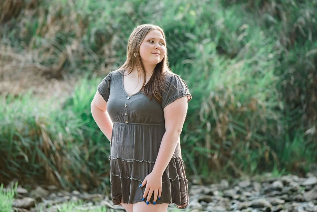 Young woman in a gray dress standing outdoors near greenery and rocks.