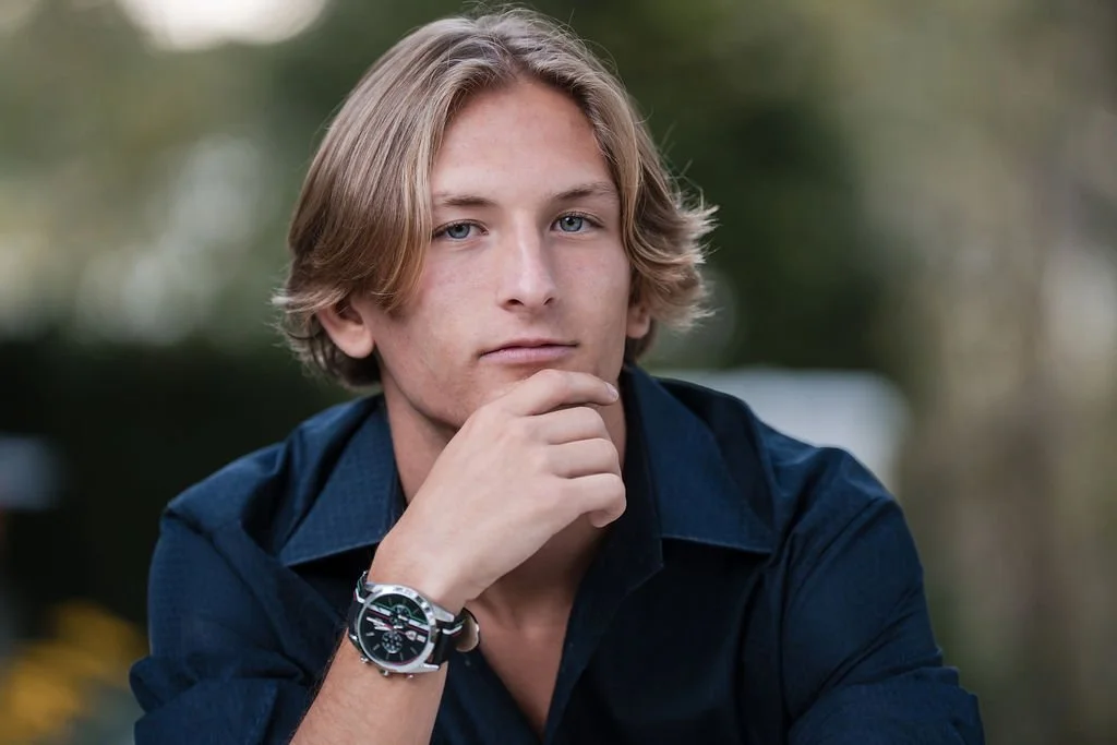 Young man with blond hair, wearing a dark blue button-up shirt and silver watch, resting his chin on his hand outdoors.