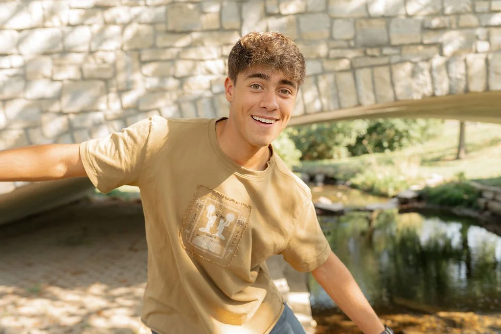 A young man smiling and posing outdoors near a small pond with a stone bridge in the background.
