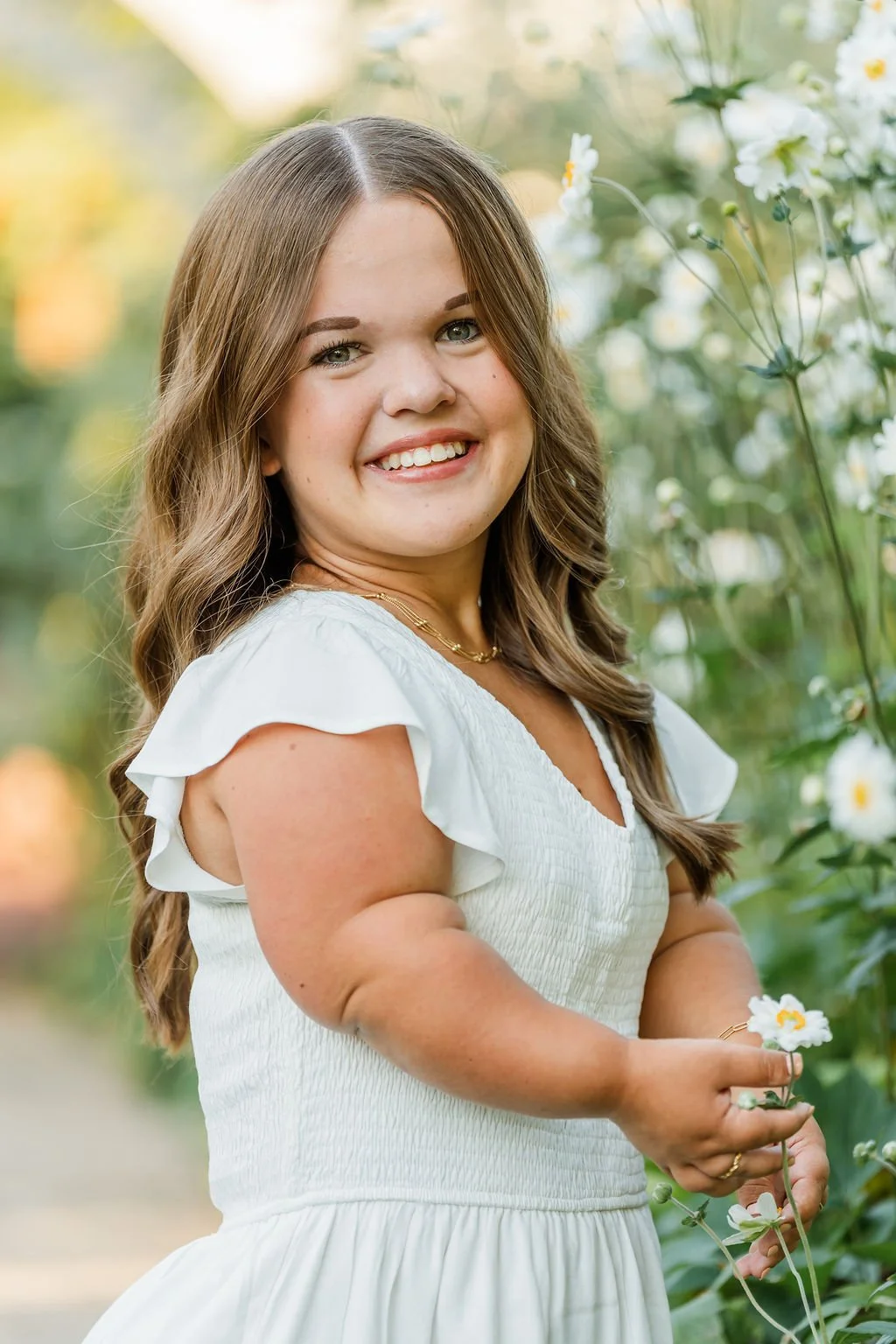 A young woman with long brown hair smiling outdoors, wearing a white dress with ruffled sleeves, holding a small white flower, with a background of green plants and flowers.